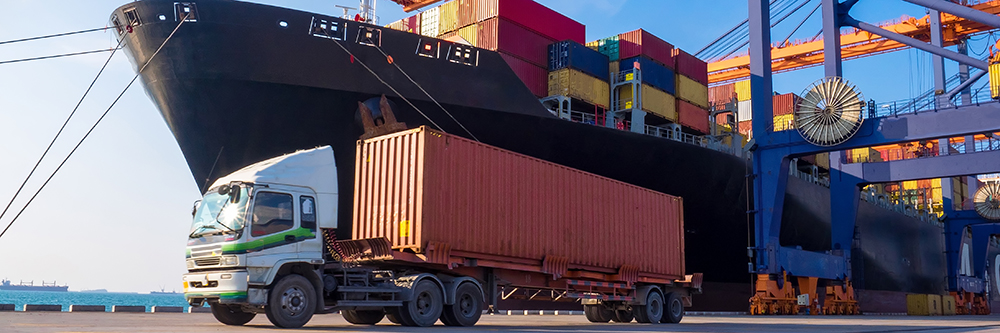 Freight truck loaded with a shipping container at a port in front of a cargo ship